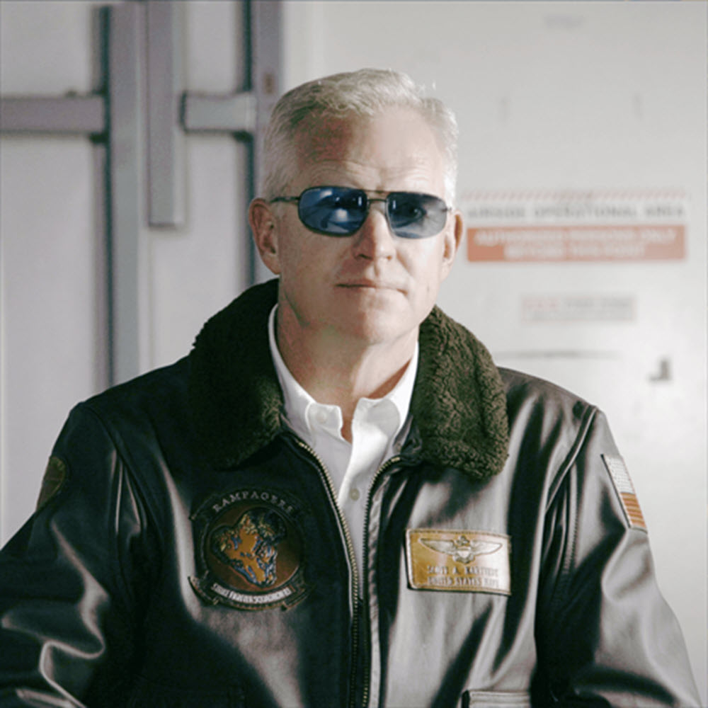 A man wearing a black leather bomber jacket with a dark green fleece collar, a white dress shirt, and Method Seven Patriot sunglasses, standing indoors against a light gray wall.
