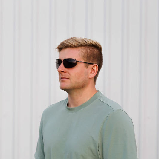 A man with blond hair wearing Method Seven Patriot aviator sunglasses and a sage green t-shirt, standing against a white corrugated metal wall.