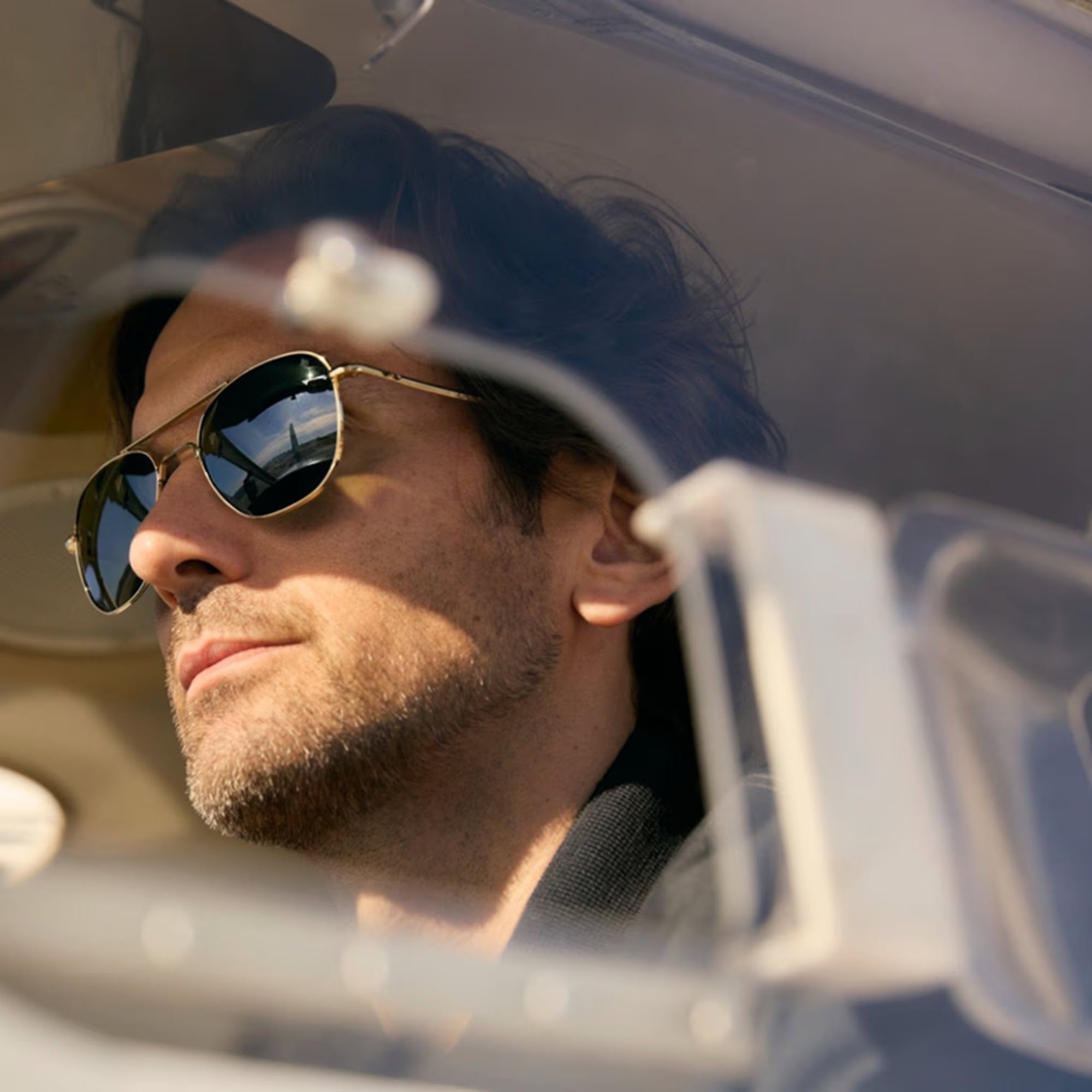 A close-up lifestyle shot of a man wearing classic aviator sunglasses inside an airplane cockpit , viewed through a side windshield.